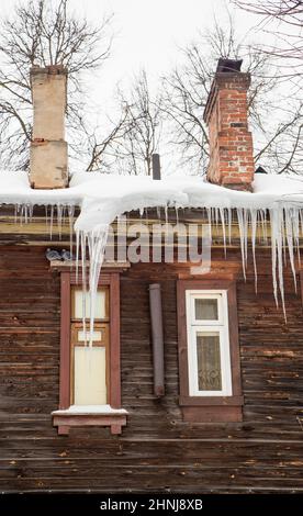 Enormi ghiaccioli trasparenti appendono sul bordo del tetto. Sullo sfondo del muro di legno della vecchia casa. Grandi cascate, anche belle file. Nuvoloso giorno d'inverno, luce tenue. Foto Stock