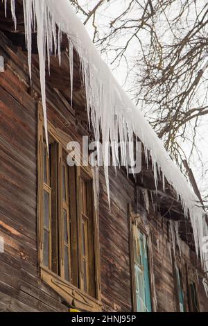 Ghiaccioli trasparenti appendono sul bordo del tetto. Sullo sfondo del muro di legno della vecchia casa. Grandi cascate, anche belle file. Nuvoloso giorno d'inverno, luce tenue. Foto Stock