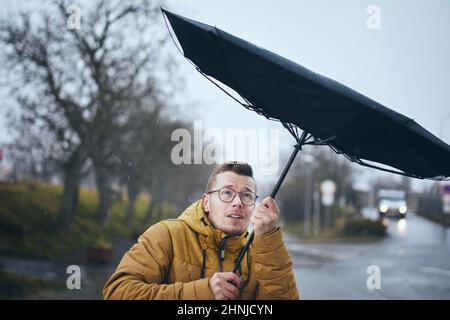 Uomo che tiene rotto ombrello in forte vento durante la notte di pioggia cupa. Temi meteo, meteorologia e cambiamenti climatici. Foto Stock
