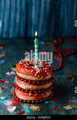 una pila di ciambelle con glassa rossa, con una candela verde e bianca illuminata sulla parte superiore, su un rustico tavolo di legno grigio cosparso di confetti di diversi co Foto Stock