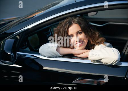 Primo piano di una signora sorridente che mette testa su braccia incrociate in cabina di auto nera. Ritratto di felice proprietario di automobile nera che riposa nella sua auto, mettendo il mento sulle braccia piegate sulla porta laterale. Foto Stock