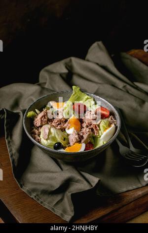Tradizionale insalata francese di nicoise con tonno in scatola, olive e uova, servita in ciotola di ceramica su tovaglia di lino scuro su tavola di legno. Rustico scuro Foto Stock