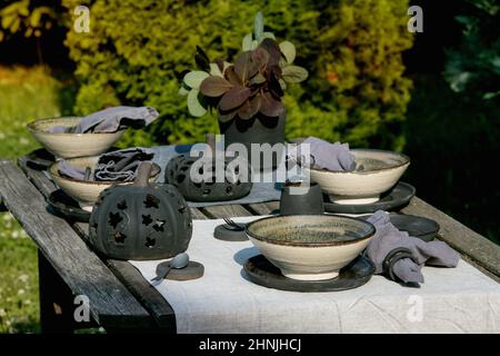 Tavola rustica ambientata all'esterno in giardino con stoviglie in ceramica artigianale vuote, piatti neri e ciotole ruvide, decorazioni in zucca, su tovaglia di lino sopra Foto Stock