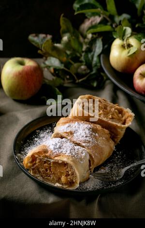 Torta tradizionale di strudel di mele a fette fatte in casa in piastra di ceramica nera servita con mele fresche mature, rami, forchetta e zucchero a velo su tavola di lino scuro Foto Stock