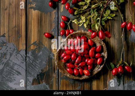 Bacche di rosa su piastra di ceramica con ramo e foglie su vecchio fondo di legno. Riserve autunnali di vitamine per l'inverno. Disposizione piatta, copia sp Foto Stock