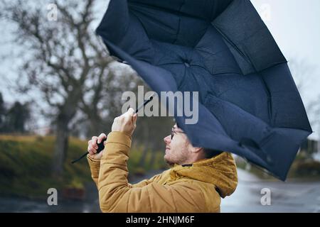 Uomo che tiene rotto ombrello in forte vento durante la notte di pioggia cupa. Temi meteo e meteorologia. Foto Stock