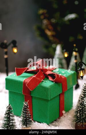 Biglietto di auguri per Natale o Capodanno. Grande scatola regalo verde con arco rosso, luci di strada giocattolo, alberi decorativi di Natale intorno su neve e abete albero a bac Foto Stock
