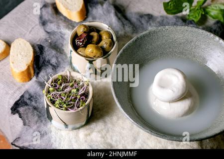Burrata tradizionale italiana annodata formaggio in ceramica grigia ciotola sul tavolo. Pane, olive, insalata verde intorno. Ingredienti per un sano din mediterraneo Foto Stock