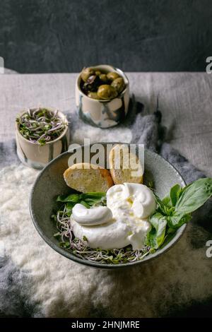Tradizionale burrata italiana annodata insalata di formaggio in ceramica grigia sul tavolo. Pane affettato, olive, germogli verdi intorno. Sana cucina mediterranea Foto Stock