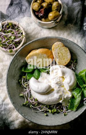 Tradizionale burrata italiana annodata insalata di formaggio in ceramica grigia sul tavolo. Pane affettato, olive, germogli verdi intorno. Sana cucina mediterranea Foto Stock