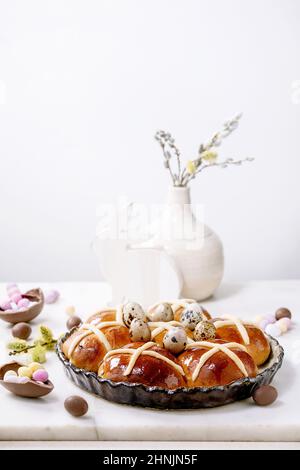 Tradizionale focaccine pasquali a croce calda fatte in casa in ceramica con fiori in fiore, caramelle al cioccolato e uova di quaglia su un tavolo di marmo bianco. Vacanze di Pasqua Foto Stock