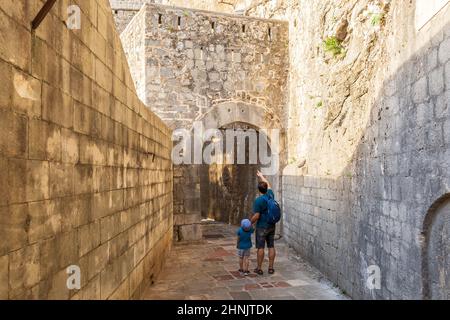 Padre e figlio camminano tra le mura medievali della fortezza nella città di Cattaro, Montenegro. I turisti esplorano la stretta strada lastricata in pietra nel centro storico su un summ Foto Stock