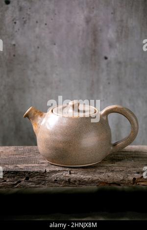 Bollitore in teiera artigianale in ceramica rextured grigio per la cerimonia del tè in piedi su un vecchio ripiano in legno con parete grigia sullo sfondo Foto Stock