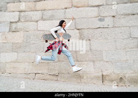 giovane ragazza hipster latino che salta intorno felice portando uno skateboard Foto Stock