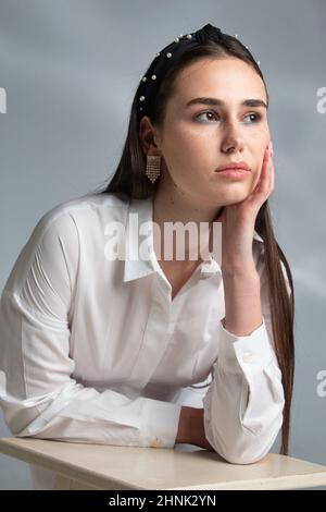 Donna in attesa con capelli scuri in blusa bianca a manica lunga appoggiata su sedia isolata in grigio Foto Stock