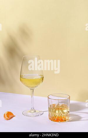 Vita morta artistica con due diversi bicchieri di vino bianco accanto a una fetta di arancio all'aperto su un tavolo bianco contro il muro giallo per spazio copia Foto Stock