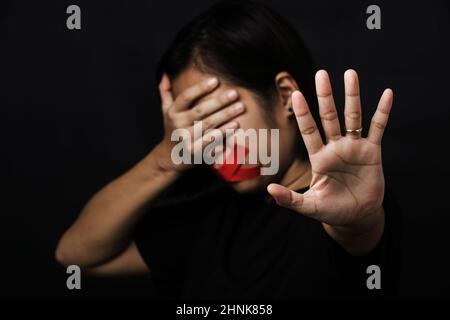 Donna bendata avvolgente bocca con nastro adesivo rosso e mostrare segno di mano smettere di abusare di violenza e abuso su sfondo nero, tratta di esseri umani e ab Foto Stock