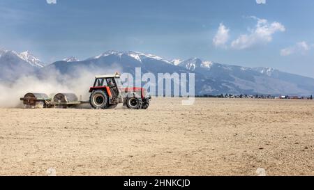 Trattore tirando heavy metal rullo su campo asciutto su un bel giorno di primavera con le montagne sullo sfondo. La preparazione del terreno. Foto Stock