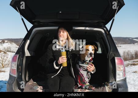 Escursioni, campeggio ed esplorazione con animali domestici: Cane in poncho e una donna in tronco SUV. Staffordshire Terrier in coperte in auto all'aperto in inverno Foto Stock