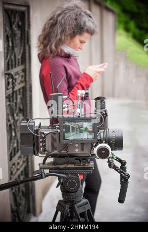 Dietro la scena. La cameraman femminile con la sua macchina fotografica tiene il telefono cellulare e chiacchiera con gli amici sul set esterno. Il videografo è in possesso di apparecchiature video Foto Stock