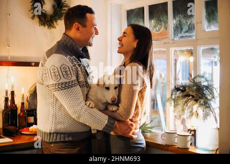 Giovane coppia felice uomo e donna tenendo carino bianco cane e abbraccio mentre in piedi in cucina decorata per Natale, bella famiglia con cucciolo godei Foto Stock