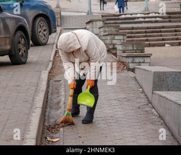 Un lavoratore con una scopa spazza la strada da polvere e detriti. Il janitor pulisce la strada. Una pulitrice femminile spazza il marciapiede con una scopa e spazzatura Foto Stock