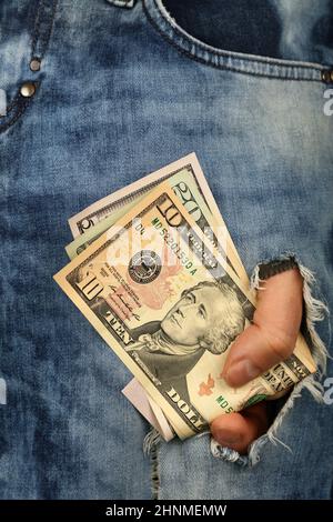 La mano tiene parecchie banconote di valuta della carta del dollaro degli Stati Uniti di valore differente in foro di lacerazione del jeans, vista ad angolo basso Foto Stock