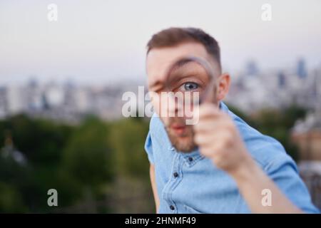 Attraente uomo closeup guardando con lente di ingrandimento alla fotocamera con sorprendente espressione facciale. Uomo che indossa una camicia jeans e un cappuccio con punta come concetto detective o investigatore. Immagine di alta qualità Foto Stock