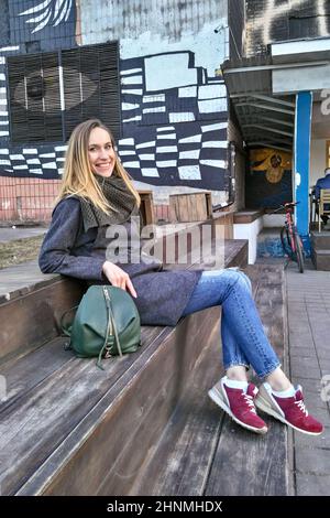 Donna fortunata in cappotto, jeans e sneakers si siede nel vecchio quartiere Foto Stock