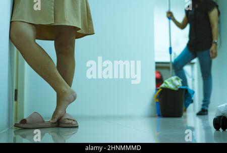 Closeup donna gamba con capelli bisogno di rimozione laser dei capelli e trattamento della pelle. Donna con gambe pelose in piedi in ufficio su una donna sfocata con mop e b Foto Stock