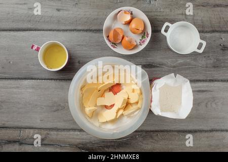 Vista del piano portapaziente - coppa in plastica con farina, tuorli d'uovo e burro, più ingredienti intorno al grigio scrivania di legno. Preparazione di torte. Foto Stock