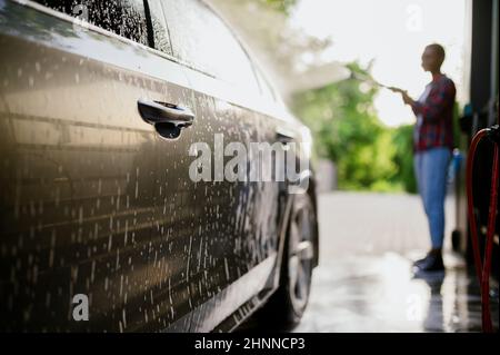 La donna lava via la schiuma dalla macchina, mano auto lavaggio stazione. Industria o commercio di autolavaggio. La persona femminile pulisce il suo veicolo dalla sporcizia all'aperto Foto Stock