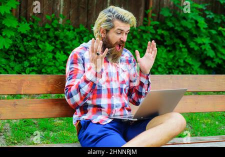 Uomo felice con il computer portatile al parco. Freelance. Gioco di soldi. Uomo barbuto con computer seduto su panca Foto Stock