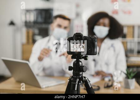 Medici multietnici maschi e femmine in maschere facciali seduti alla scrivania e utilizzando la macchina fotografica moderna per filmare video vlog. Consigli online durante la pandemia da parte di personale medico competente. Foto Stock