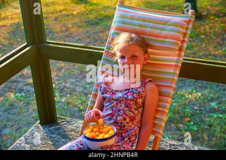 La bambina è seduta su una sedia a sdraio da giardino su una veranda. Piccola ragazza è mangiare formaggio aromatizzati snack in giardino. Sognante e immagine romantica. Estate e infanzia felice concetto Foto Stock