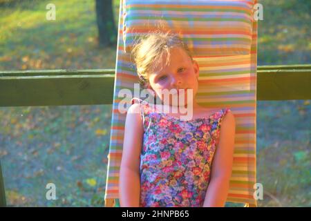 La bambina è seduta su una sedia a sdraio da giardino su una veranda. Piccola ragazza sta prendendo il sole in giardino. Sognante e immagine romantica. Estate e infanzia felice concetto Foto Stock