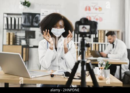 Medico qualificato africano femmina che mostra come indossare maschera medica viso durante la quarantena durante la registrazione video vlog in sala d'ospedale. Concetto di telemedicina, persone e tecnologia. Foto Stock