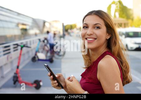 Primo piano di una giovane donna d'affari sicura si gira e sorride con l'app per smartphone si sta preparando a utilizzare lo scooter elettrico per tornare a casa Foto Stock