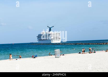 Persone che si godono la spiaggia accanto a una nave da crociera che lascia il porto di Miami per i Caraibi Foto Stock