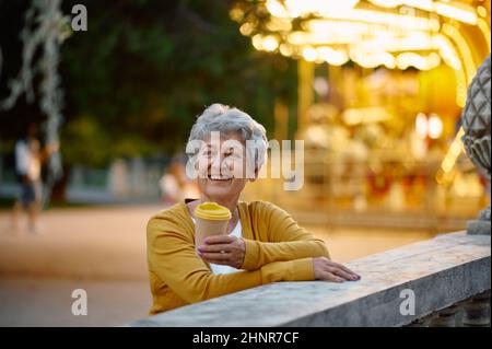 Grazioso caffè Granny drink nel parco divertimenti estivo, illuminazione luminosa sullo sfondo. Stile di vita delle persone anziane. Divertente nonna che si diverte all'aria aperta, Foto Stock