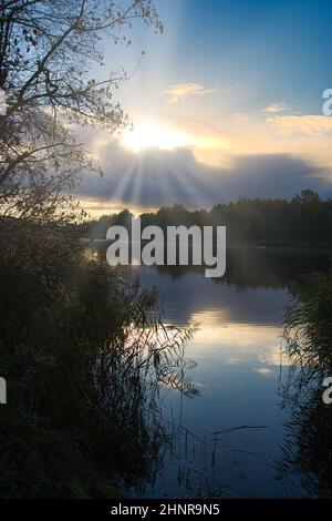 Sunburst attraverso le nuvole con fasci di luce che cadono su un lago tranquillo sotto circondato da verdi foreste verdeggianti vista attraverso un gap tra gli alberi Foto Stock