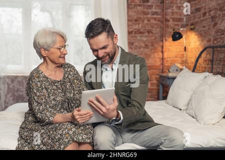 signora caucasica anziana dai capelli grigi godendo il suo tempo e guardando le foto sul tablet del suo figlio di mezza età. Foto di alta qualità Foto Stock