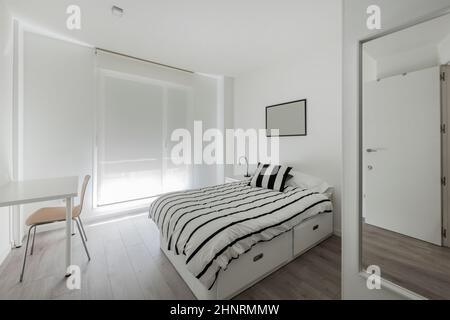 Camera decorata in tonalità bianche con un letto con cassetti in legno e un piumone a righe blu Foto Stock