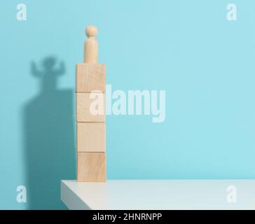 la statuetta di legno di un uomo si erge in alto sui cubetti, l'ombra di un forte e vittorioso. Tutti sono più forti di quanto pensi, un supereroe. Dorso blu Foto Stock