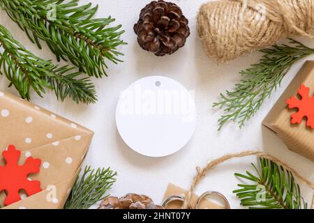 Etichetta regalo di carta rotonda di Natale su un tavolo bianco con rami di abete, regali avvolti e decorazioni per le vacanze vista dall'alto. Rustica composizione invernale w Foto Stock