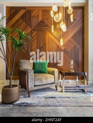 Moderna poltrona beige, tavolino da caffè, piantatrice, alto lampadario di vetro moderno e pareti rivestite in legno decorate Foto Stock