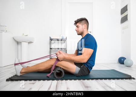 Giovane uomo che usa la cintura di Yoga mentre fa l'esercitazione sul mat di idoneità alla clinica di riabilitazione. Foto Stock