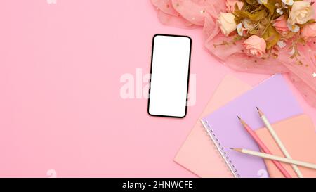 Tavolo da studio femminile o scrivania da ufficio con schermo bianco per smartphone, notebook colorati, matite, bouquet di rose e spazio per la copia su sfondo rosa Foto Stock