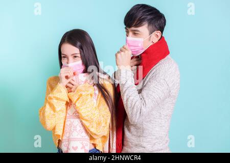 Giovane coppia che indossa sciarpe e maschere mediche, facendo gesti freddi e tosse. Isolato su sfondo blu Foto Stock