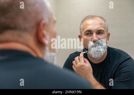 Uomo maturo dai capelli grigi che restyling la sua barba in casa utilizzando il rasoio Foto Stock